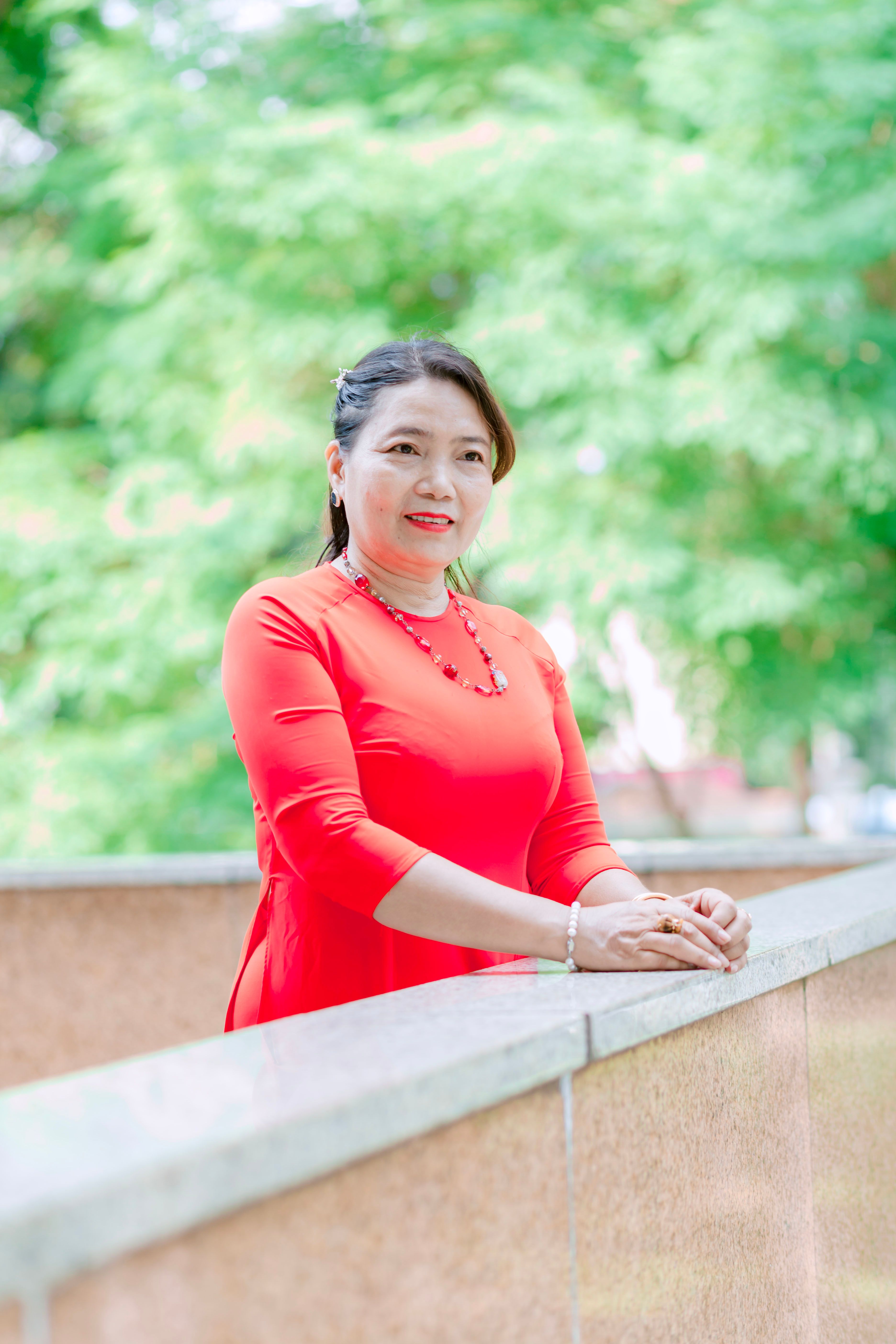 Asian woman in vibrant red dress standing by a park bridge in sunlight.
