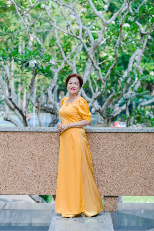 Free Asian woman in a yellow dress posing outdoors with lush green trees in the background. Stock Photo
