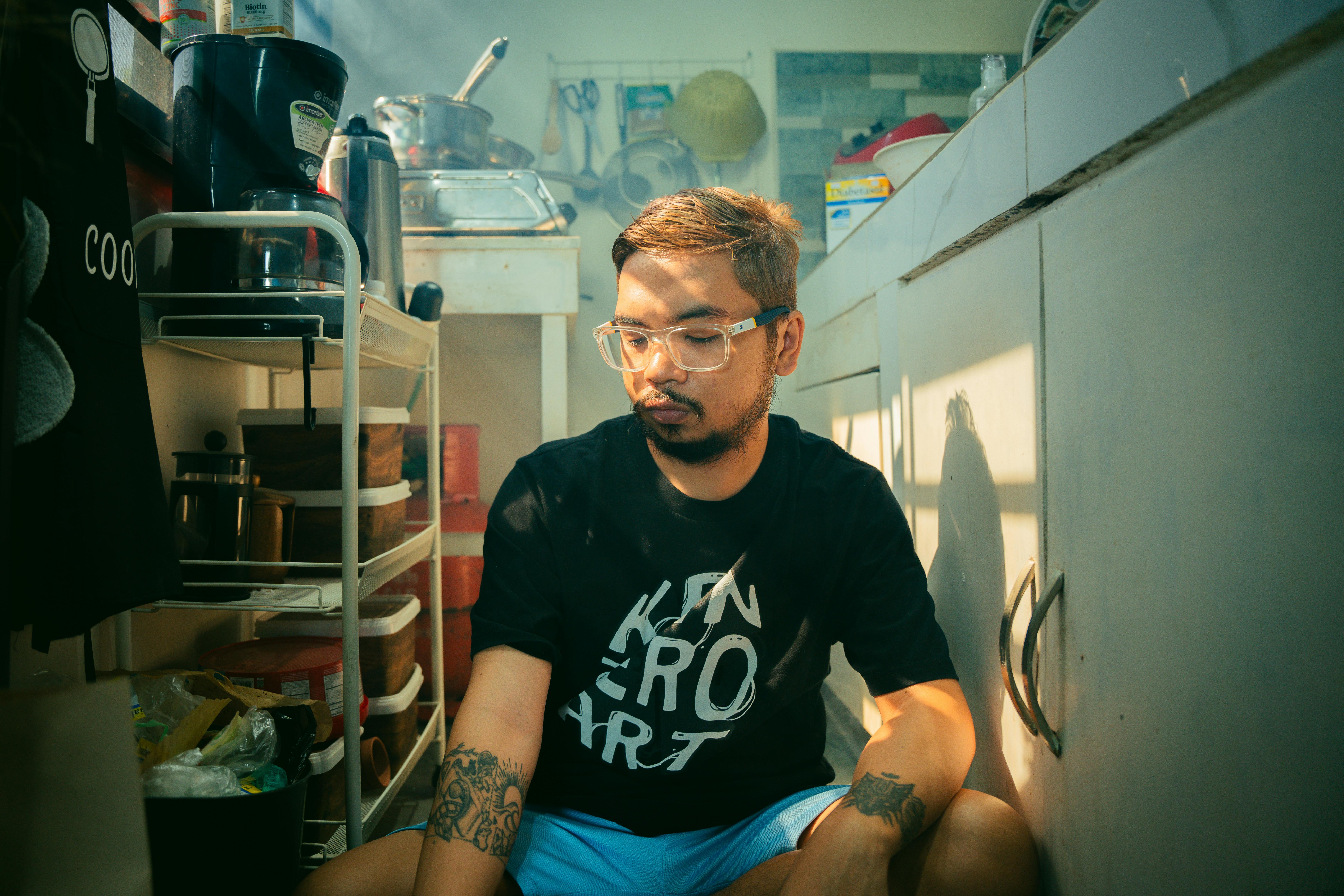 Man with eyeglasses sitting in a sunlit kitchen, surrounded by kitchenware, wearing a graphic t-shirt.
