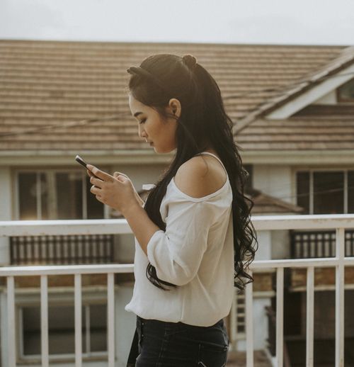Free Side profile of a young woman with long hair using a smartphone on a balcony. Stock Photo