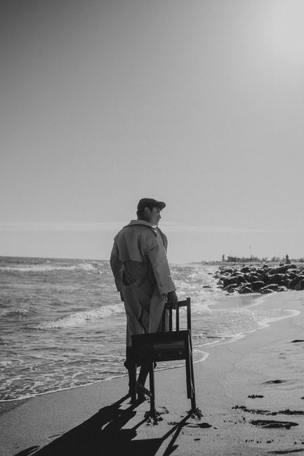 A man stands on Barcelona's beach holding a chair, captured in dramatic black and white.