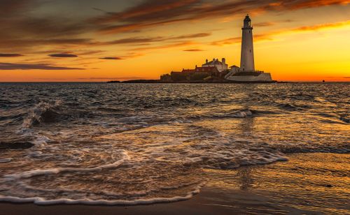 Free Stunning sunset behind St Mary's Lighthouse with waves crashing on the shore, creating a serene coastal view. Stock Photo
