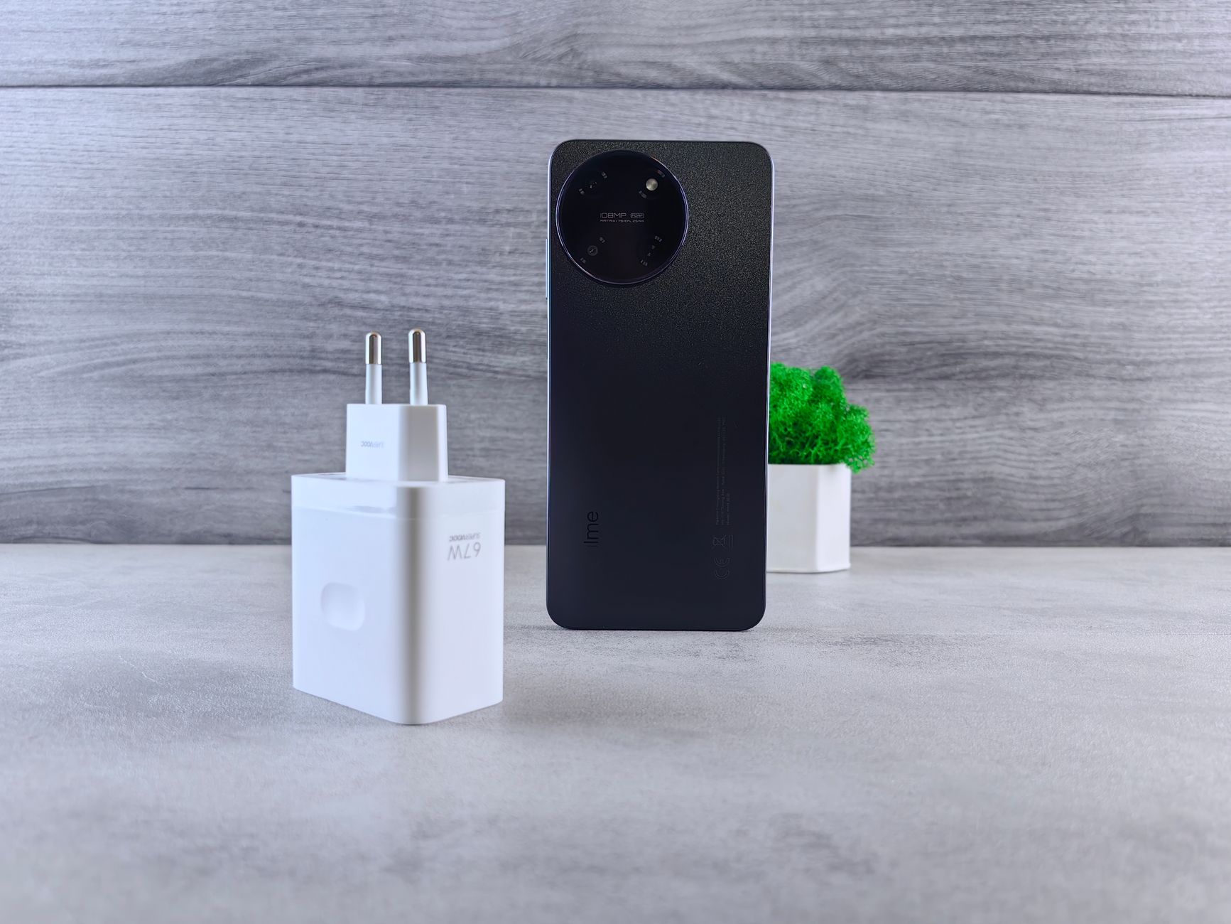Minimalist composition of a smartphone, charger, and small green plant on a grey background.