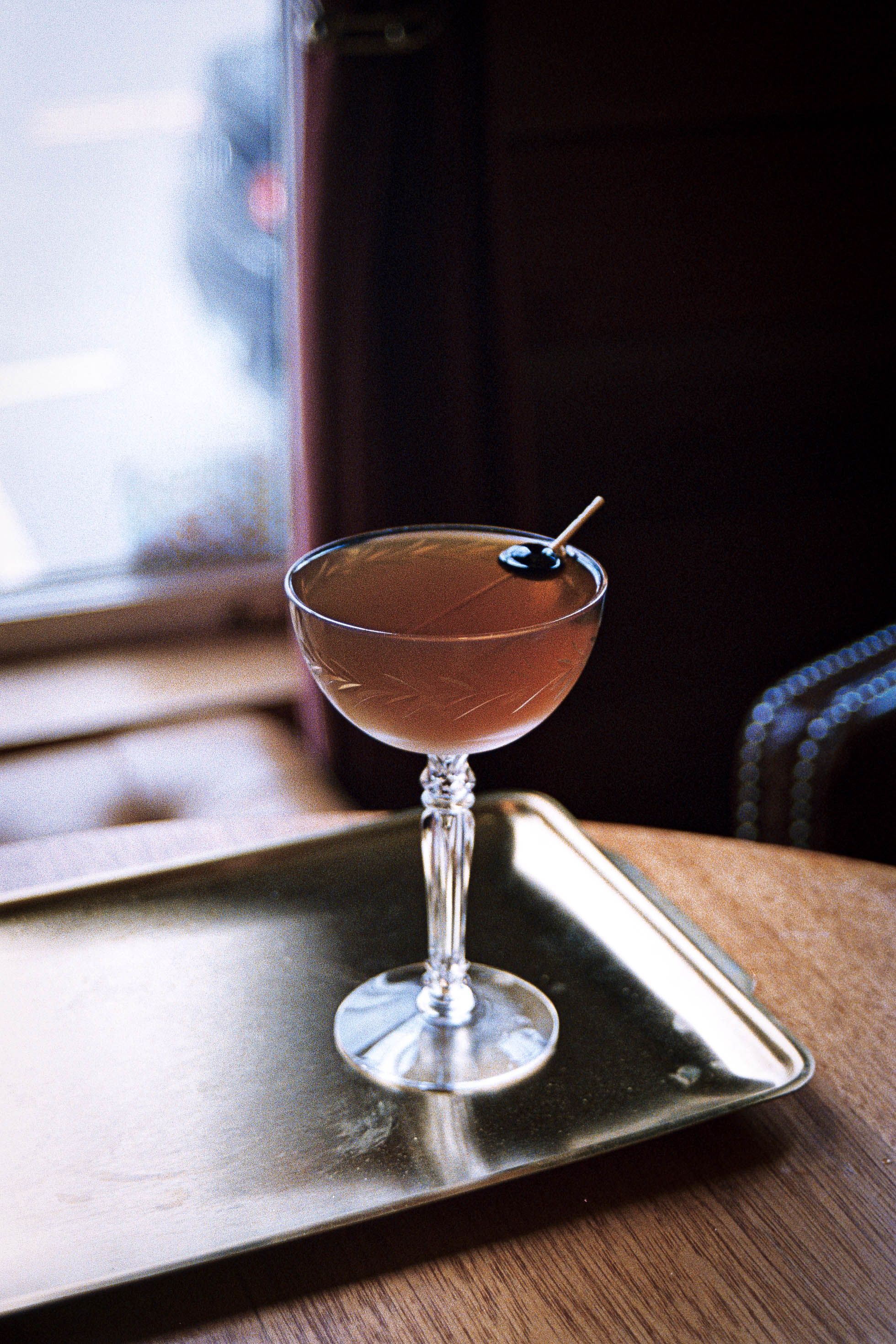 A refined cocktail in a crystal glass sits on a golden tray by a window, capturing elegance.