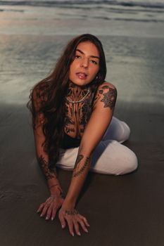 A tattooed woman poses elegantly on a beach at dusk, exuding style and sensuality.