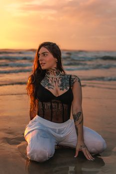 A tattooed woman kneels on the beach at sunset, combining beauty and art in a striking fashion photograph.