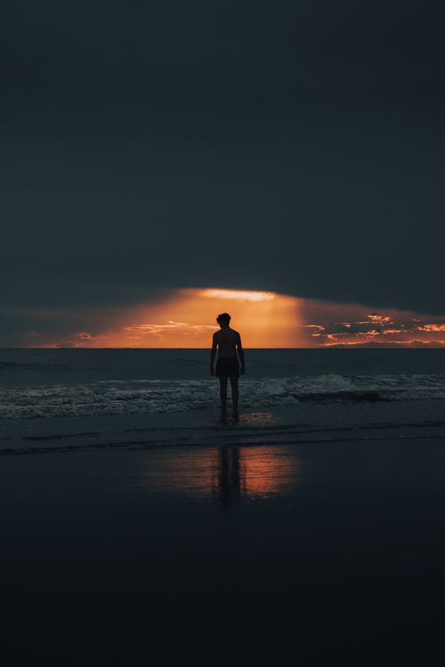 Free Silhouette of a man standing on the beach during a dramatic sunset with dark clouds and radiant light. Stock Photo