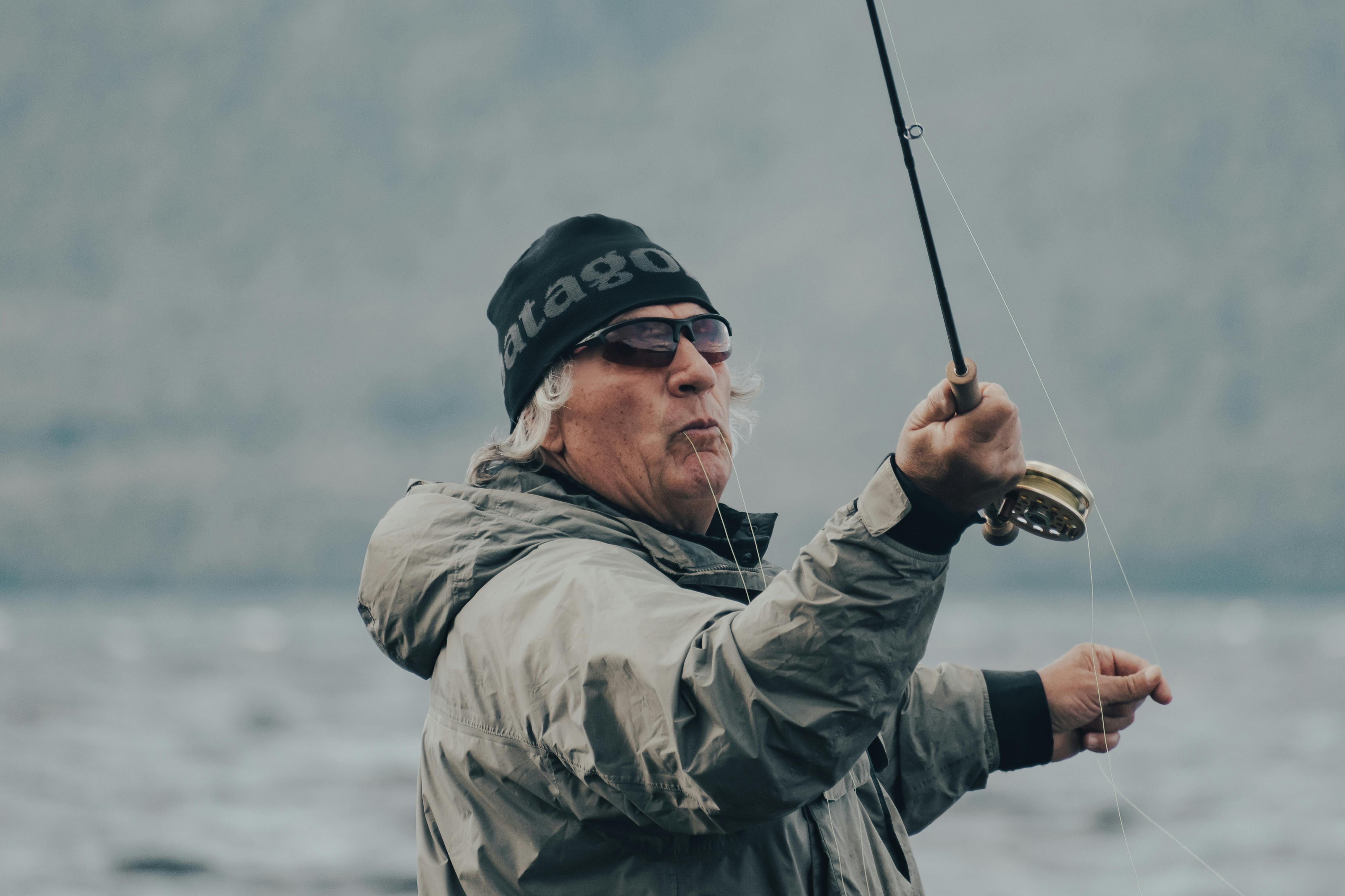 A senior man in a jacket and beanie fly fishing by the sea on a cloudy day.