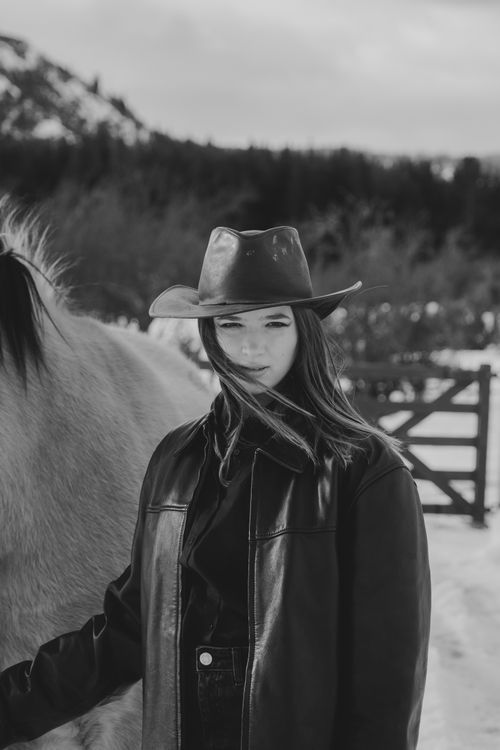 Free A woman in a cowboy hat and leather jacket stands beside a horse in a snowy landscape. Stock Photo