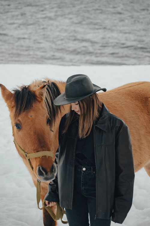 Free Woman in leather jacket stands with brown horse on snowy shore during winter. Stock Photo