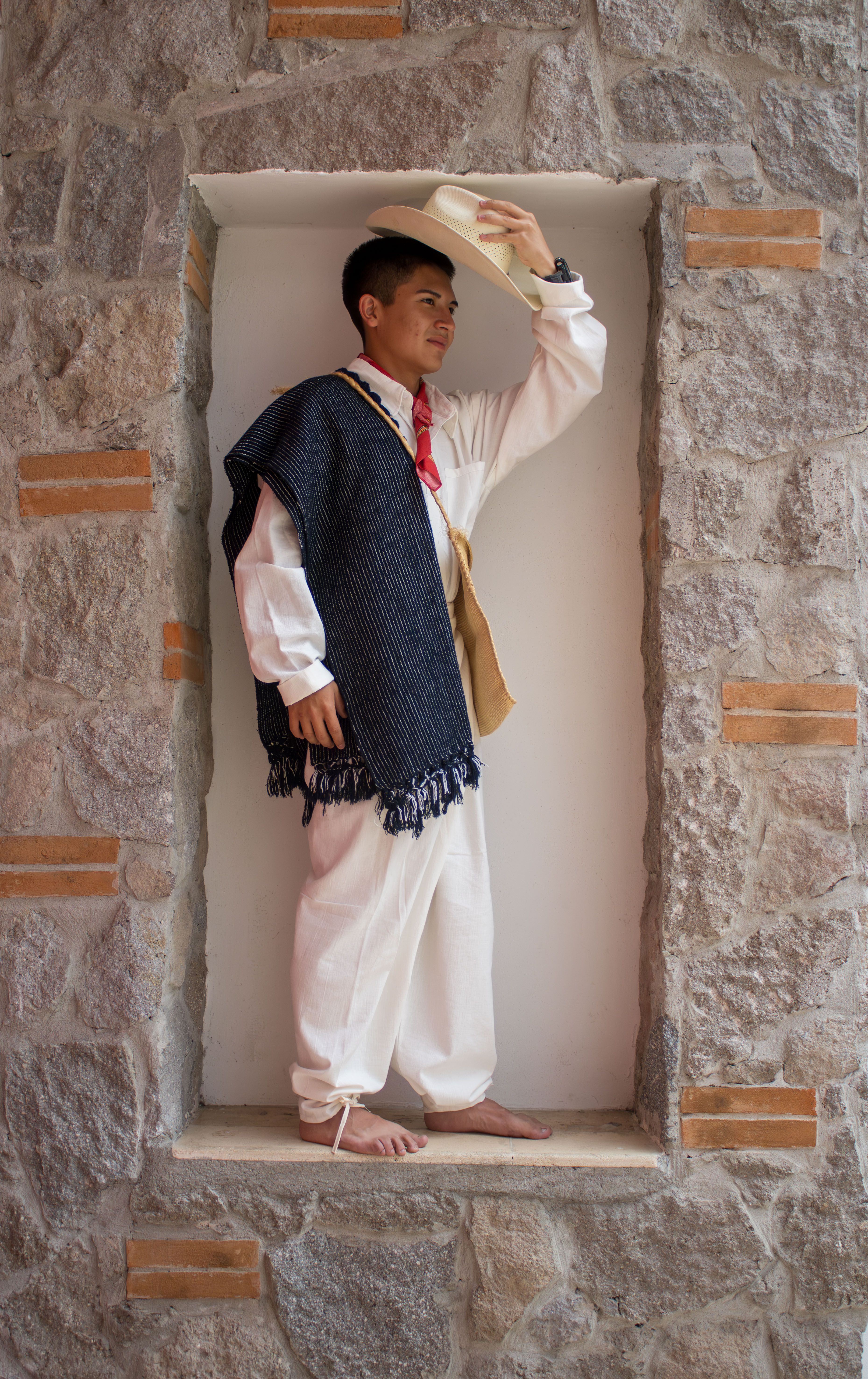 A young man wearing traditional clothing and a hat poses barefoot in a stone wall niche.