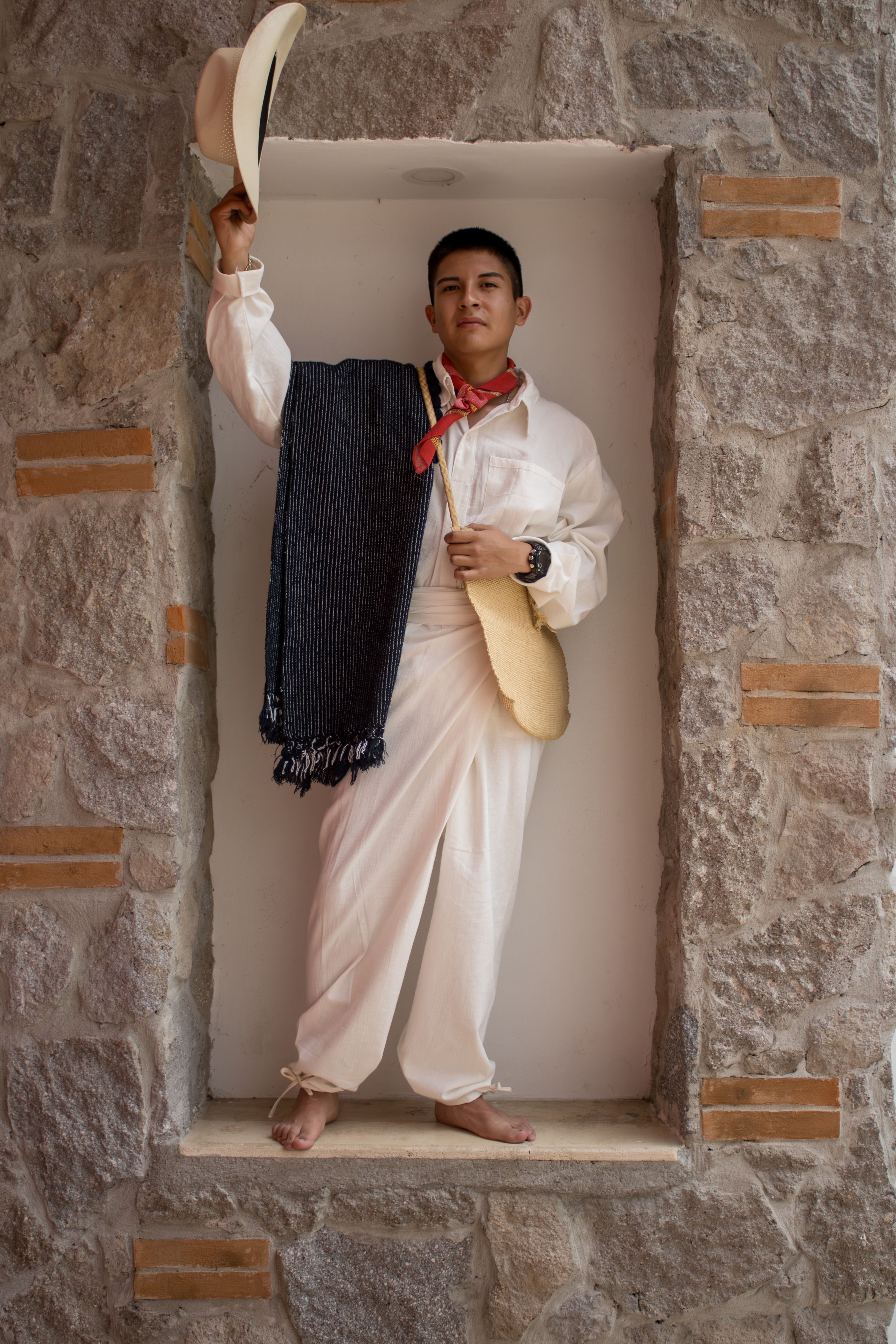 Man in traditional clothing poses barefoot in a stone recess, holding a hat.