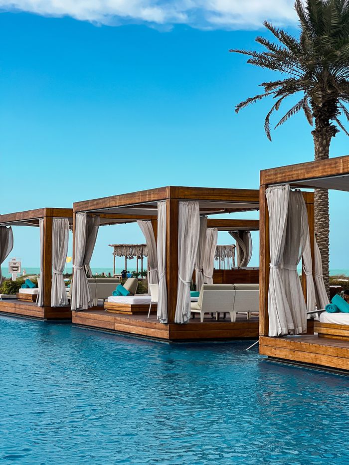 Poolside cabanas with white drapes standing in turquoise water under a palm