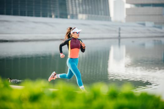 Athletic woman jogging in vibrant sportswear by an urban lakeside setting during the day.