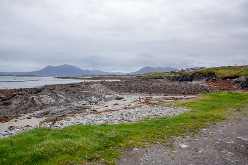 Free Tranquil Irish coast with rocky shore, grassy path, and distant mountains under cloudy skies. Stock Photo