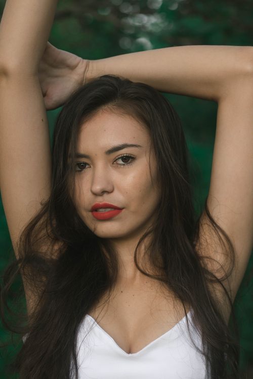 Free Young gorgeous ethnic lady with long dark hair and red lips standing against green plants with hands behind head and looking at camera Stock Photo