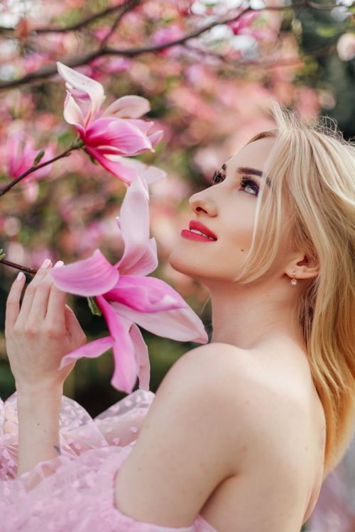 Free A captivating portrait of a woman with blonde hair admiring pink magnolia blossoms outdoors in spring. Stock Photo