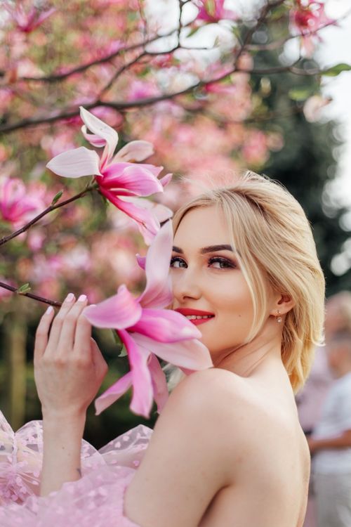 Free A blonde woman in a floral dress smiles near blooming pink blossoms during spring. Stock Photo