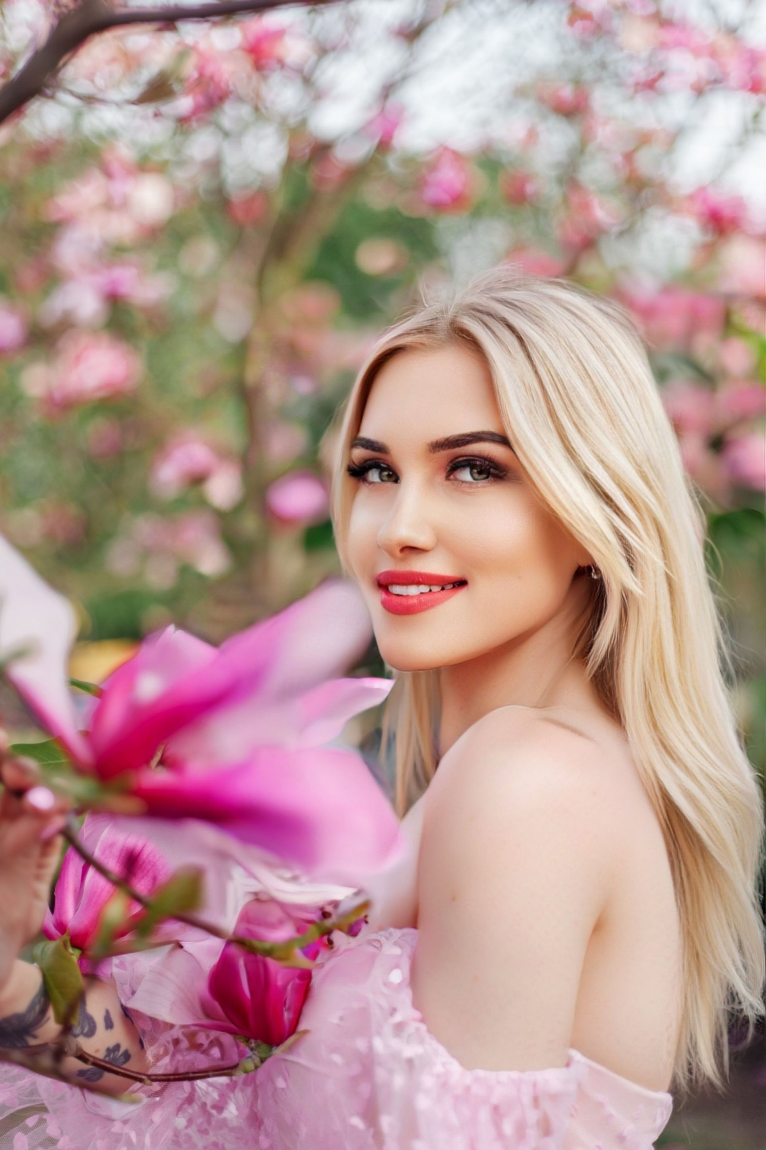 A blonde woman smiles amidst vibrant pink blossoms in the spring outdoors.