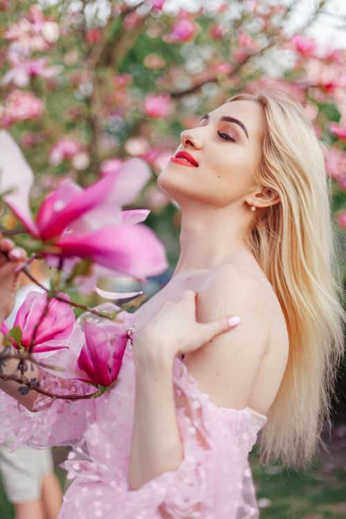 Free Blonde woman wearing pink dress posing gracefully with magnolia flowers in spring. Stock Photo