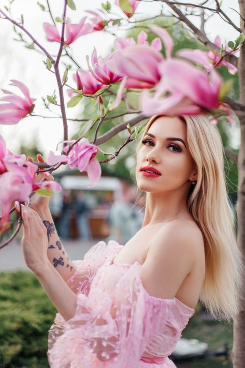 Free A beautiful blonde woman poses gracefully with blooming magnolia flowers in a stylish outdoor portrait. Stock Photo