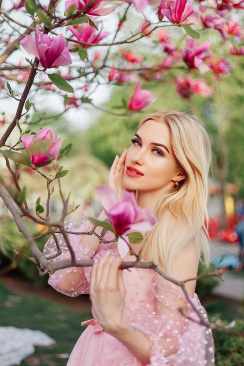 Free A blonde woman in a pink dress poses among blooming magnolia trees, capturing the essence of spring fashion. Stock Photo