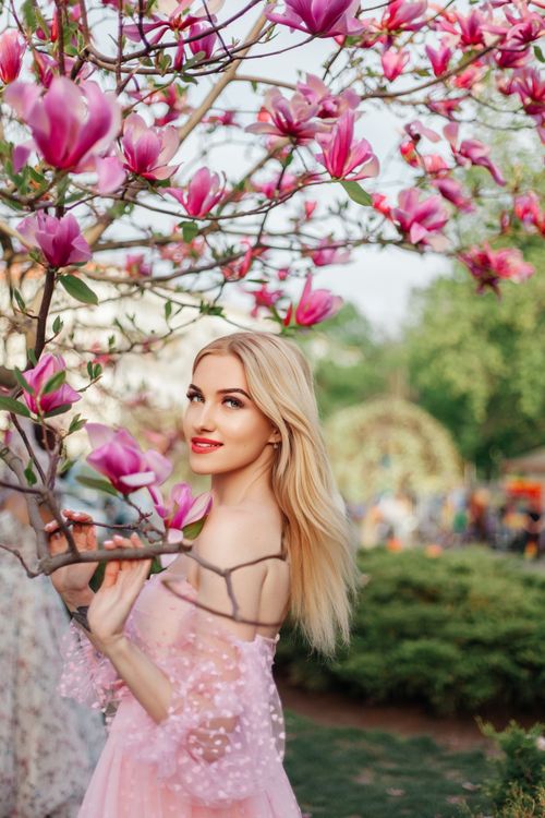 Free Elegant blonde woman in pink dress smiling beside blooming magnolia tree in spring garden. Stock Photo