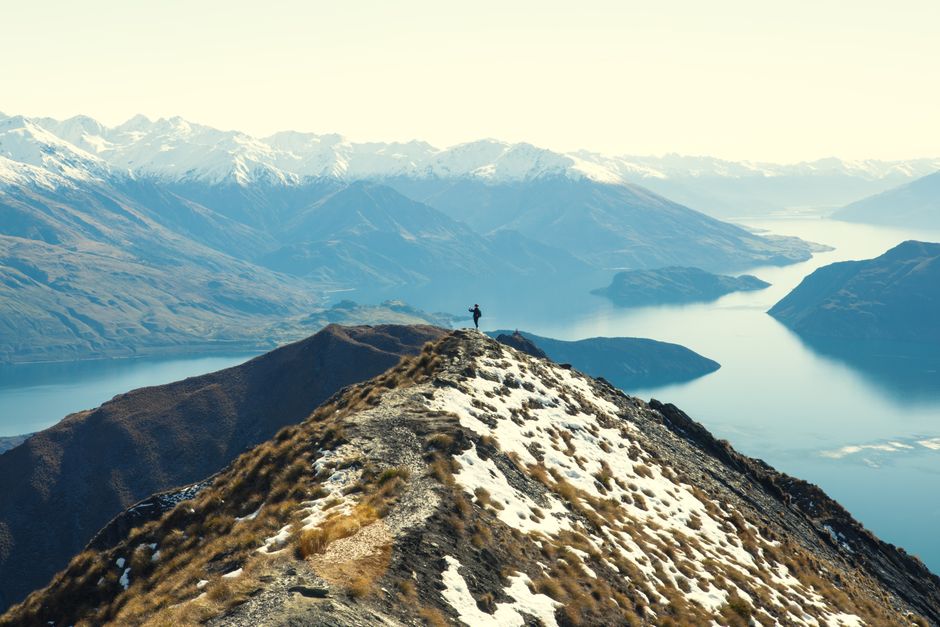 A lone hiker stands on a stunning snowy ridge with majestic mountains and a pristine lake view.