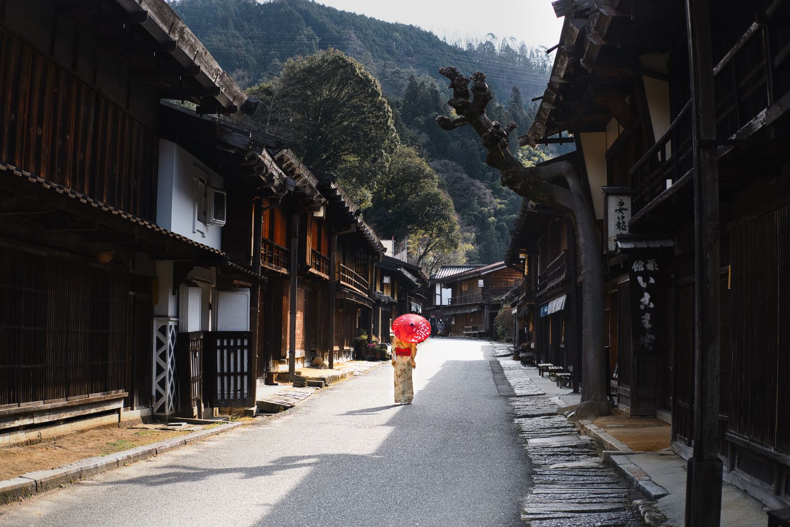 A woman with a red parasol walks the empty street of a preserved post town on the old Nakasendo road, dark wooden inns beneath a forested mountain in Japan's Kiso Valley