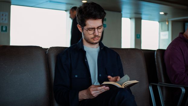 Focused young man with glasses reads a book while sitting indoors. Quiet and serene atmosphere.