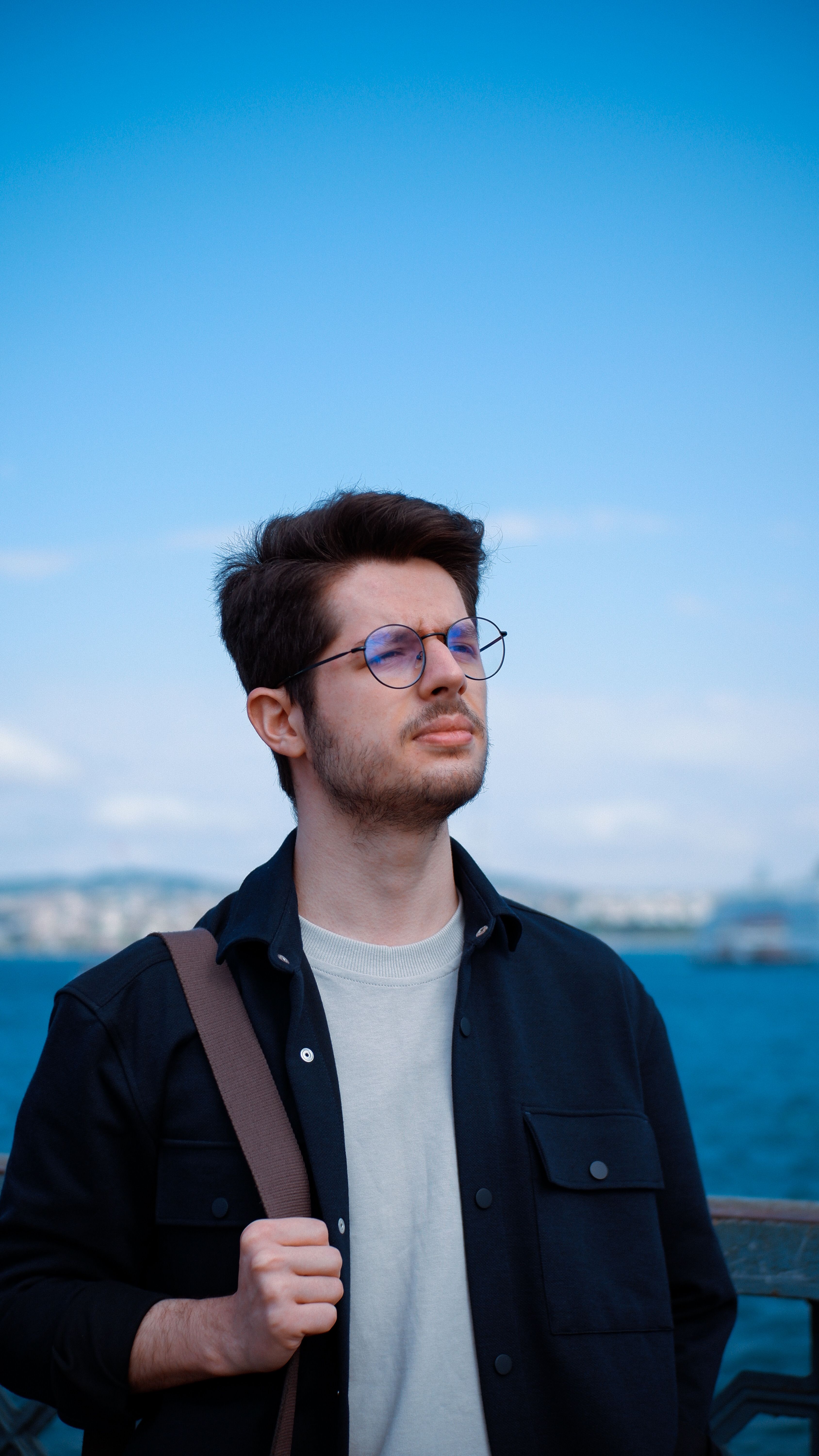 Portrait of a stylish man wearing glasses by the waterfront under a bright blue sky.