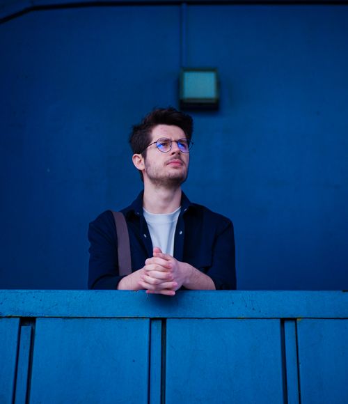 Free A young man with eyeglasses stands thoughtfully against a blue wall, exuding a contemplative mood. Stock Photo