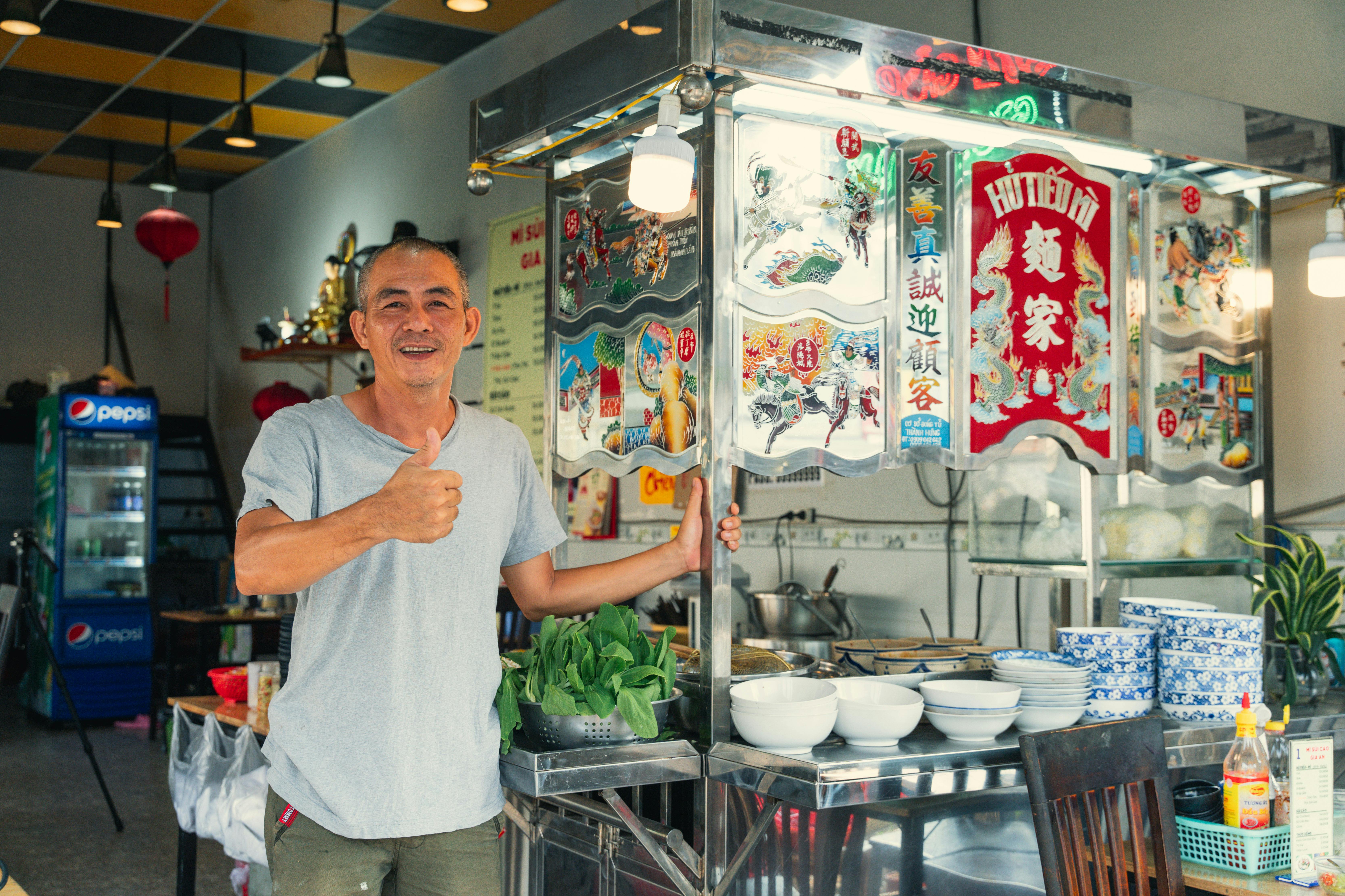 Smiling man giving a thumbs up at a vibrant market stall in Ho Chi Minh City, Vietnam.