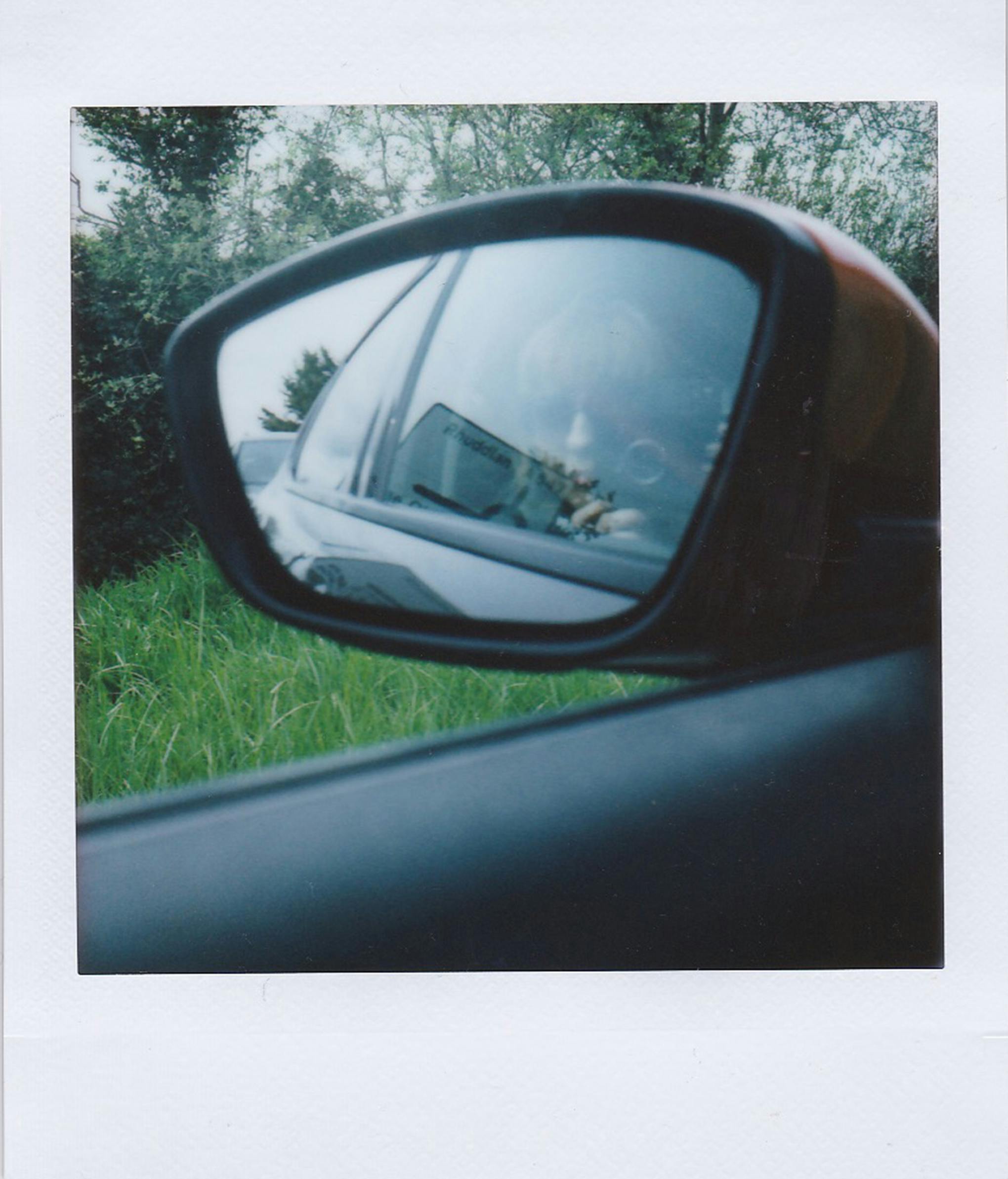 Rearview mirror capturing a reflection of a person inside a parked car by the roadside.