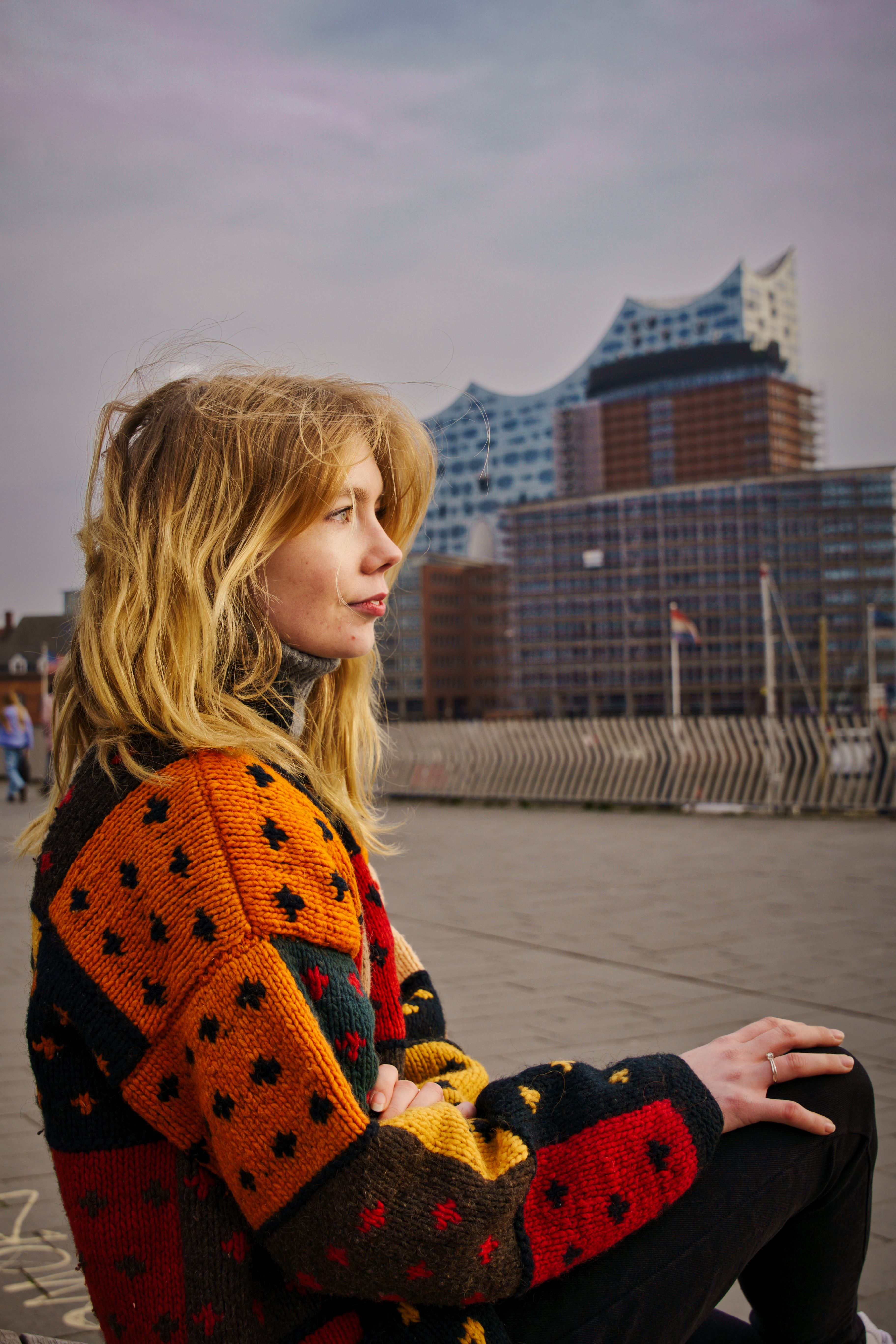 A young woman in a colorful sweater poses outdoors near the Elbphilharmonie in Hamburg, Germany.