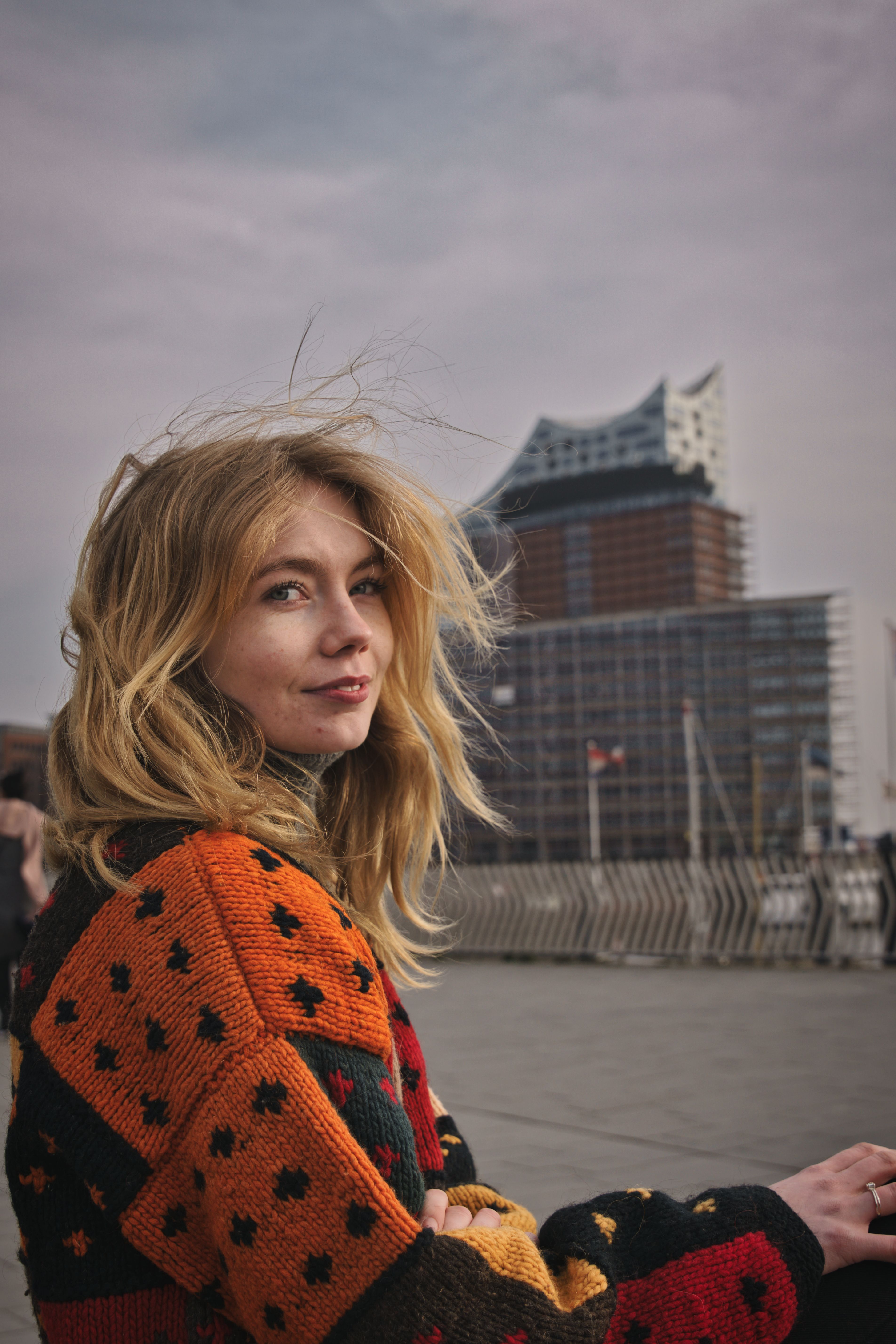 Young woman in colorful sweater sitting outdoors with Elbphilharmonie Hamburg in the background.