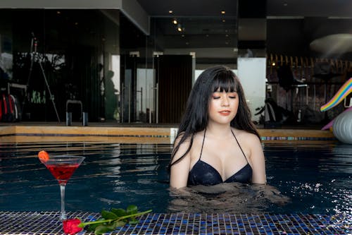 女人在游泳池附近的葡萄酒和红玫瑰花朵杯上穿着黑色比基尼上衣