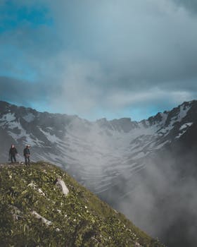两名徒步旅行者穿越雾蒙蒙的山地，天空壮阔，山峰白雪皑皑