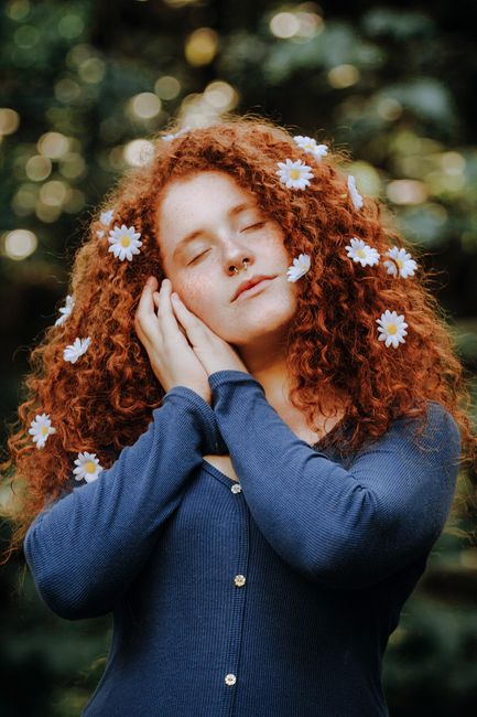A serene young woman with curly red hair adorned with daisy flowers, eyes closed, in a natural setting.