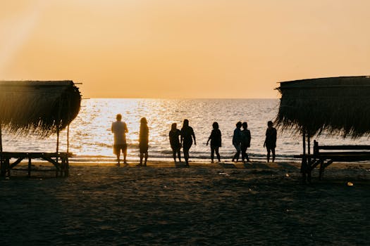 People silhouetted against a golden beach sunset with thatched huts in view, creating a warm and serene atmosphere.