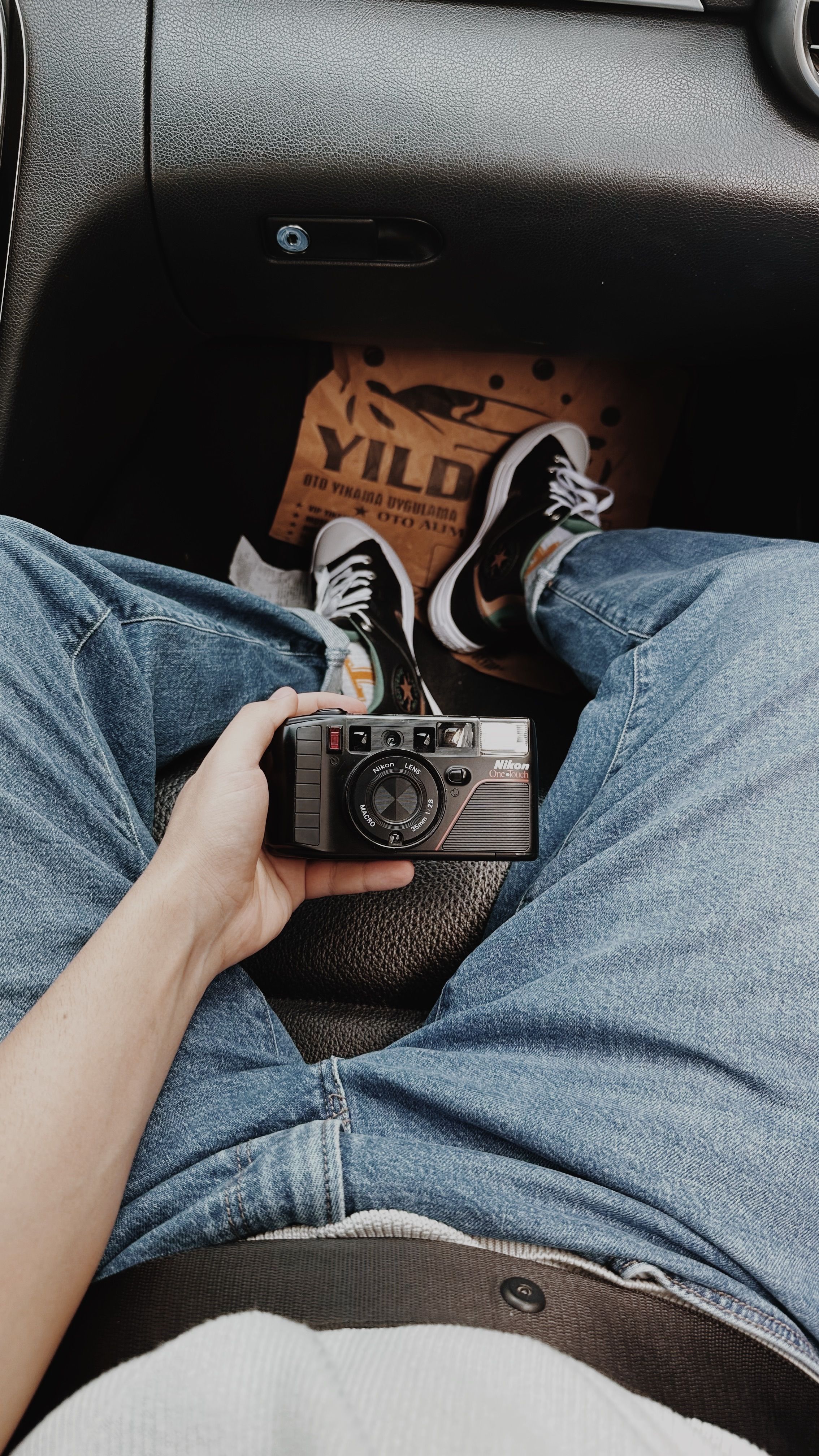 Close-up of person holding a vintage camera while sitting in a car interior wearing jeans and sneakers.