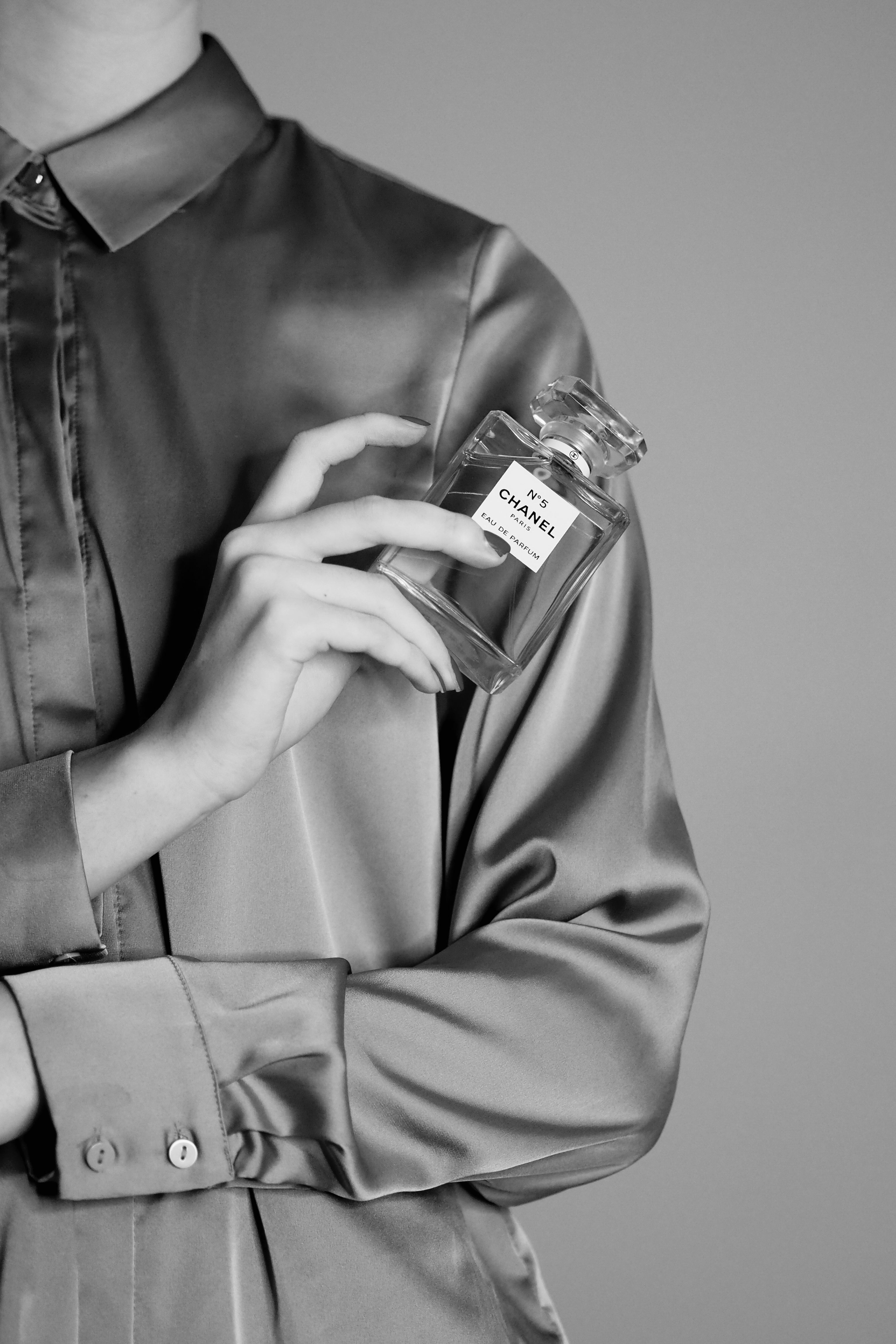 Black and white image of a person holding a Chanel perfume bottle against a satin shirt background.
