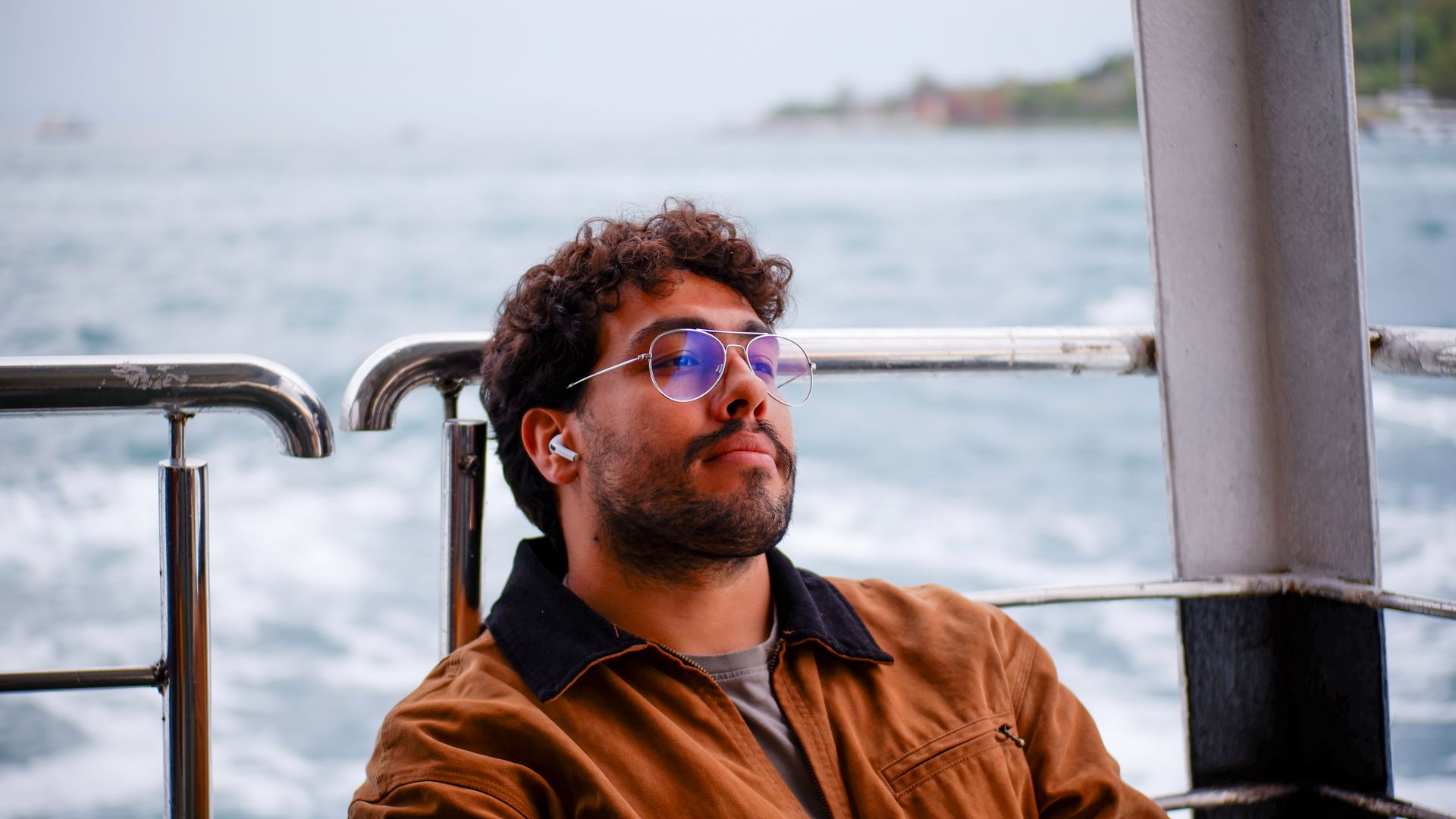 Man relaxing on a ferry, wearing wireless earbuds and sunglasses, with a sea view.
