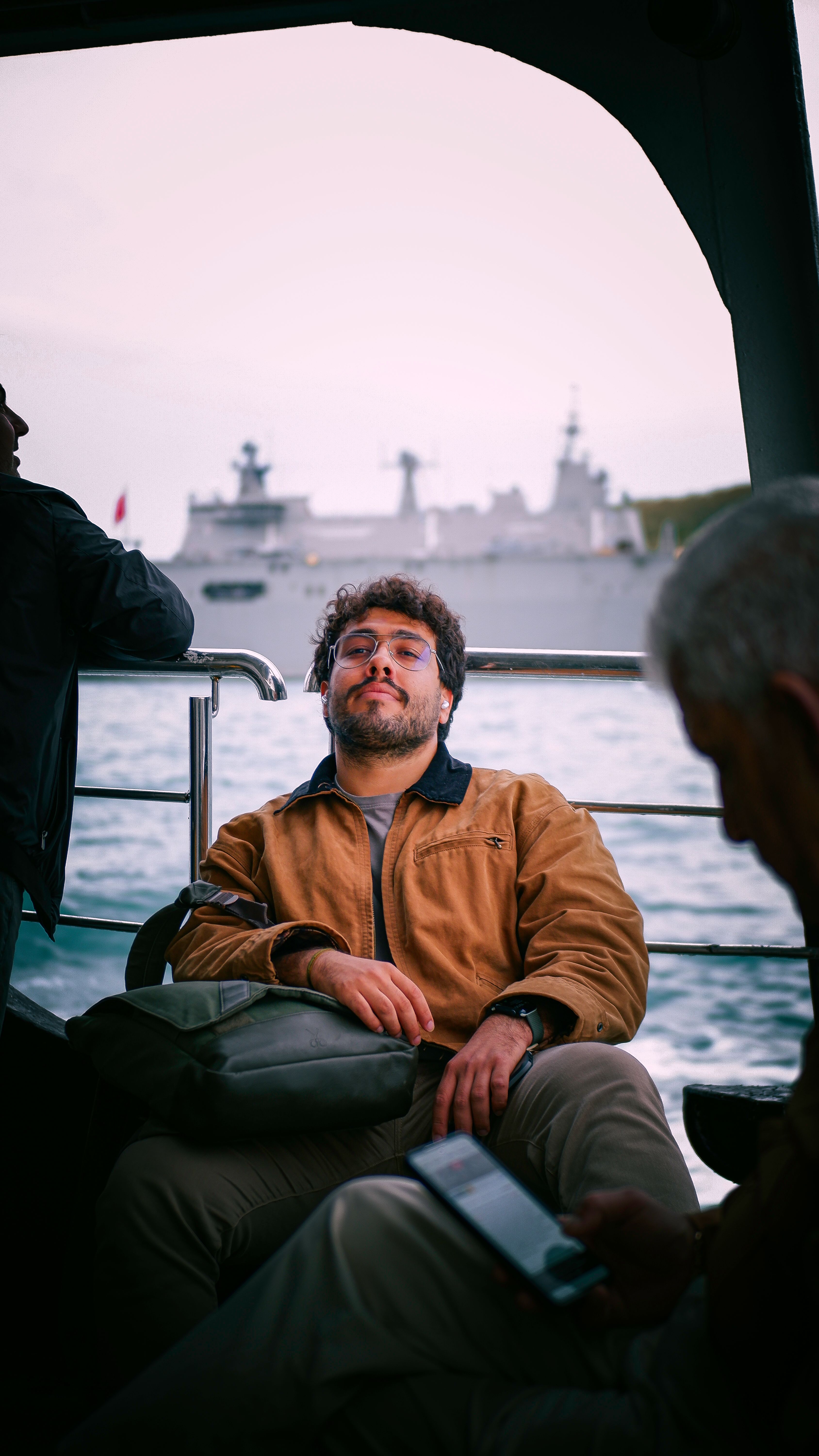 A man sitting on a ferry, gazing at the sea with a ship in the background.