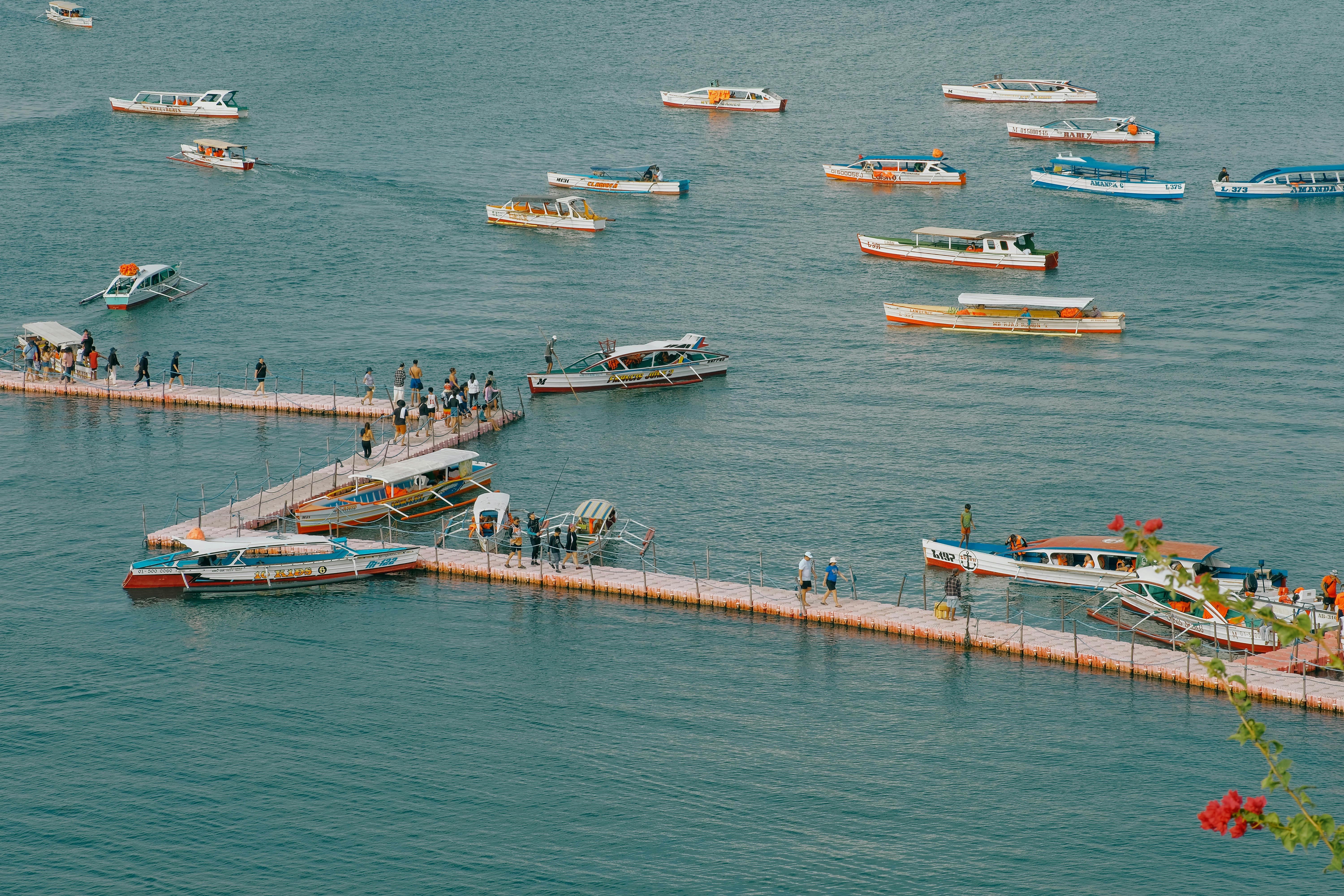 A vibrant harbor scene featuring numerous boats docked along a zigzagging floating pier, teeming with people, set against a peaceful sea backdrop.