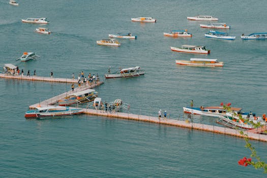 A vibrant harbor scene featuring numerous boats docked along a zigzagging floating pier, teeming with people, set against a peaceful sea backdrop.