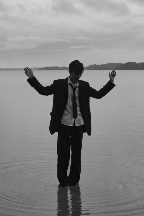 Free Black and white photo of an Asian man in a suit standing with arms raised on a lakeshore. Stock Photo