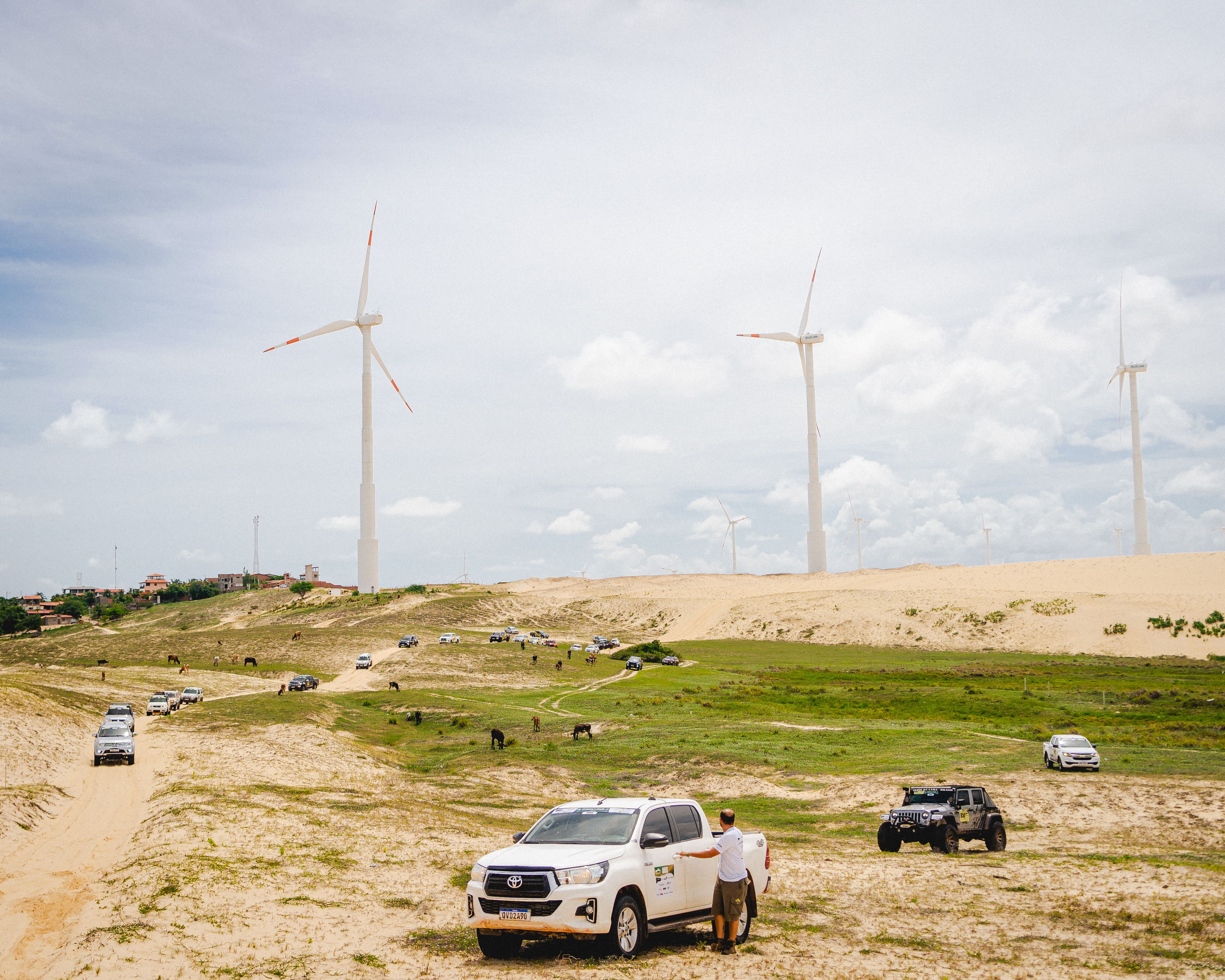 Wide shot of cars traversing sandy landscape with wind turbines in the background, showcasing renewable energy and adventure.