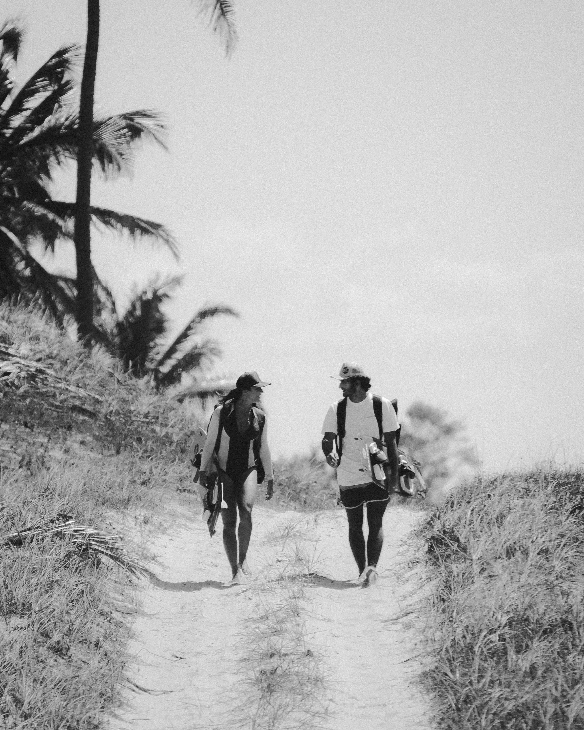 A couple enjoys a leisurely walk on a sandy seaside path amidst palm trees.