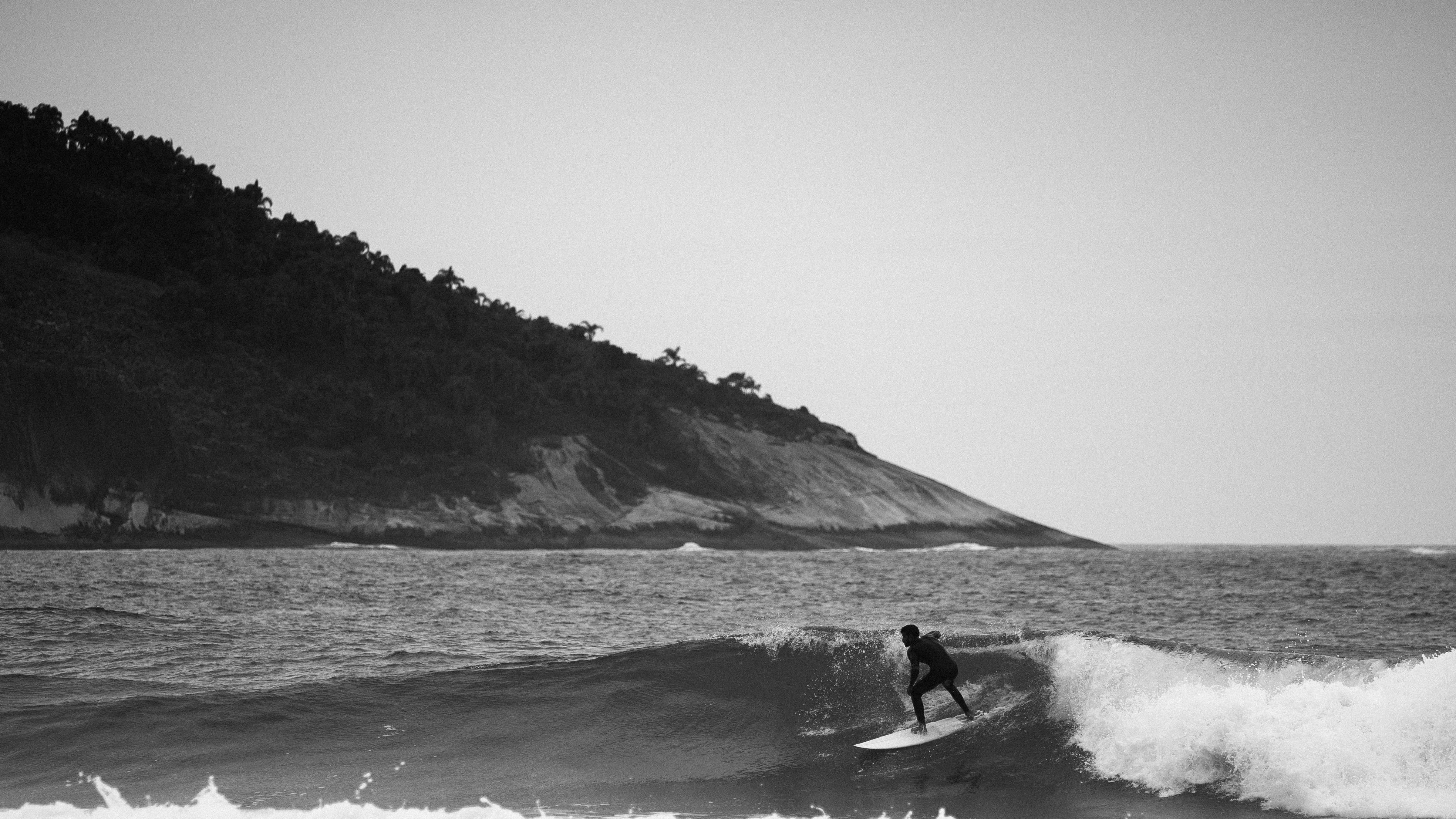 Monochrome image of a surfer catching waves near a rugged coastline.